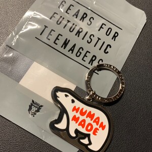 May include: A silver key chain with a white polar bear charm. The charm has the words "HUMAN MADE" printed in red on it. The key chain is packaged in a plastic bag with the words "GEARS FOR FUTURISTIC TEENAGERS" printed on it.