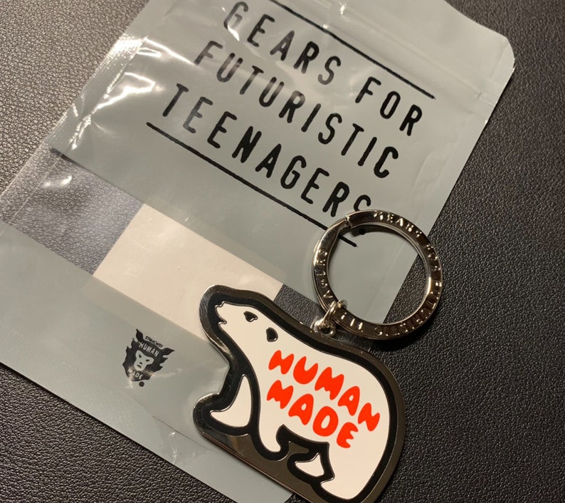 May include: A silver key chain with a white polar bear charm. The charm has the words "HUMAN MADE" printed in red on it. The key chain is packaged in a plastic bag with the words "GEARS FOR FUTURISTIC TEENAGERS" printed on it.