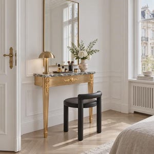 Op de afbeelding: Een gouden sidetable met een grijs marmeren blad, een rechthoekige spiegel met een gouden lijst, een gouden paddenstoelvormige lamp en een zwarte kruk met een gebogen rugleuning. De tafel heeft sierlijke gouden houtsnijwerken op de poten en lade.