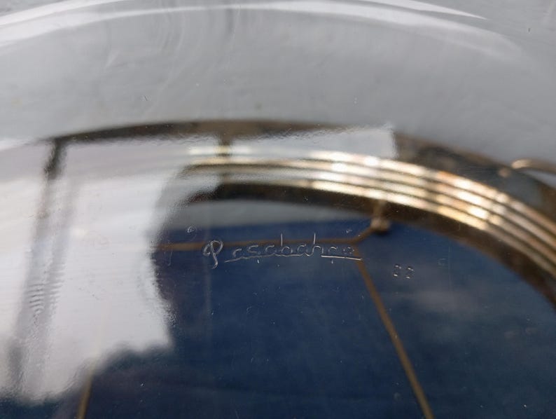May include: A clear glass cake stand with a gold metal base. The base has three tiers and the glass top is etched with the word "Pessence".