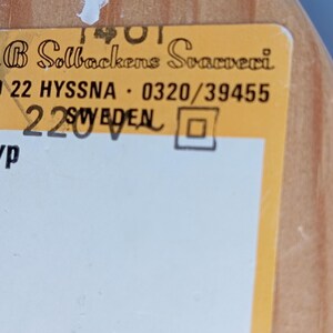 Op de afbeelding: Een geel label met zwarte tekst luidt "G B Solbackens Svamperi" en "510 22 HYSSNA 0320/39455 220V SWEDEN". Het label heeft ook het woord "Typ" en een vierkant.