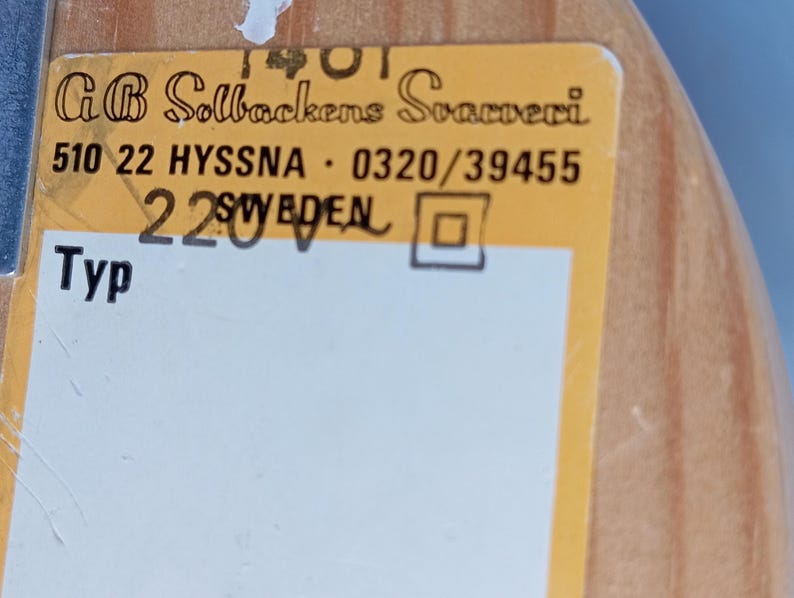 Op de afbeelding: Een geel label met zwarte tekst luidt "G B Solbackens Svamperi" en "510 22 HYSSNA 0320/39455 220V SWEDEN". Het label heeft ook het woord "Typ" en een vierkant.