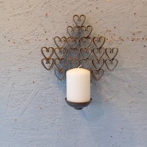May include: A metal wall sconce with a heart-shaped design. The sconce features a series of interconnected hearts arranged in a triangular pattern. A white pillar candle sits in a dark metal holder at the base of the design.