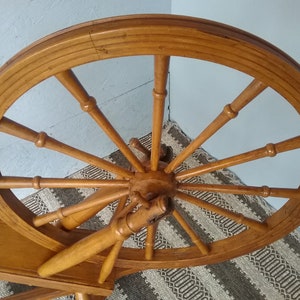 May include: A large, circular wooden spinning wheel with twelve spokes radiating from a central hub. The wheel features a smooth, light brown finish and is connected to a horizontal wooden frame, showcasing a classic design with turned wood details.