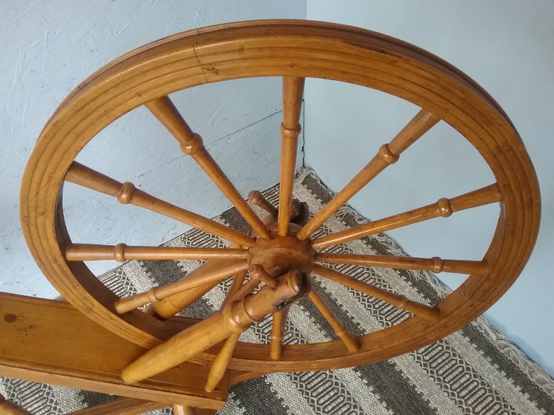 May include: A large, circular wooden spinning wheel with twelve spokes radiating from a central hub. The wheel features a smooth, light brown finish and is connected to a horizontal wooden frame, showcasing a classic design with turned wood details.