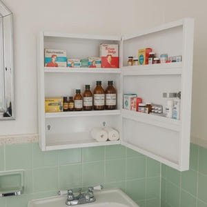 May include: A white medicine cabinet with the door open, revealing shelves stocked with various vintage medicine bottles and boxes. The cabinet is mounted on a wall above a sink and green tile.