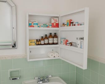 May include: A white medicine cabinet with the door open, revealing shelves stocked with various vintage medicine bottles and boxes. The cabinet is mounted on a wall above a sink and green tile.