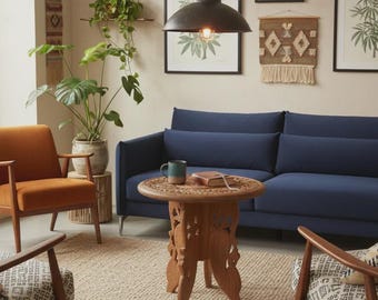 May include: A living room featuring a navy blue sofa, an orange armchair, and two patterned chairs. A round, carved wooden side table sits in the center with a mug and notebook. A black pendant light hangs above, with botanical art on the wall.