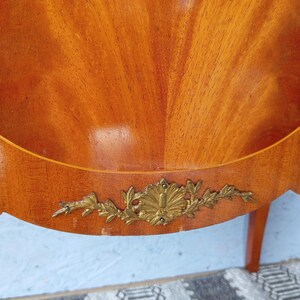 May include: Close-up of a wooden furniture piece with a warm, reddish-brown finish. The wood grain is prominently displayed in a fan-like pattern. A decorative gold-colored metal embellishment is affixed to the lower edge, adding a touch of elegance.
