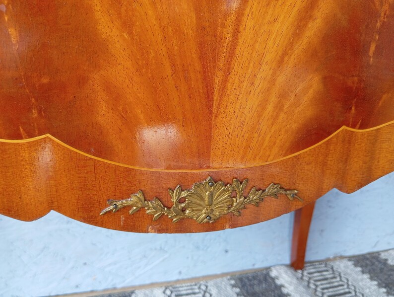 May include: Close-up of a wooden furniture piece with a warm, reddish-brown finish. The wood grain is prominently displayed in a fan-like pattern. A decorative gold-colored metal embellishment is affixed to the lower edge, adding a touch of elegance.