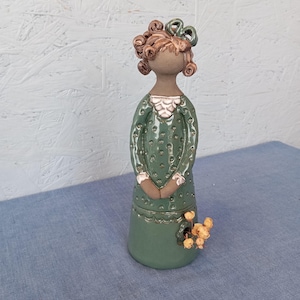 May include: A ceramic figurine of a woman with brown hair in a green polka dot dress. She is holding a bouquet of yellow flowers.