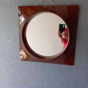May include: A square wall mirror with a glossy dark brown frame and a circular center glass. The mirror features a simple rope hanger attached to the top edge, allowing it to be mounted securely on a wall surface for display and reflection.