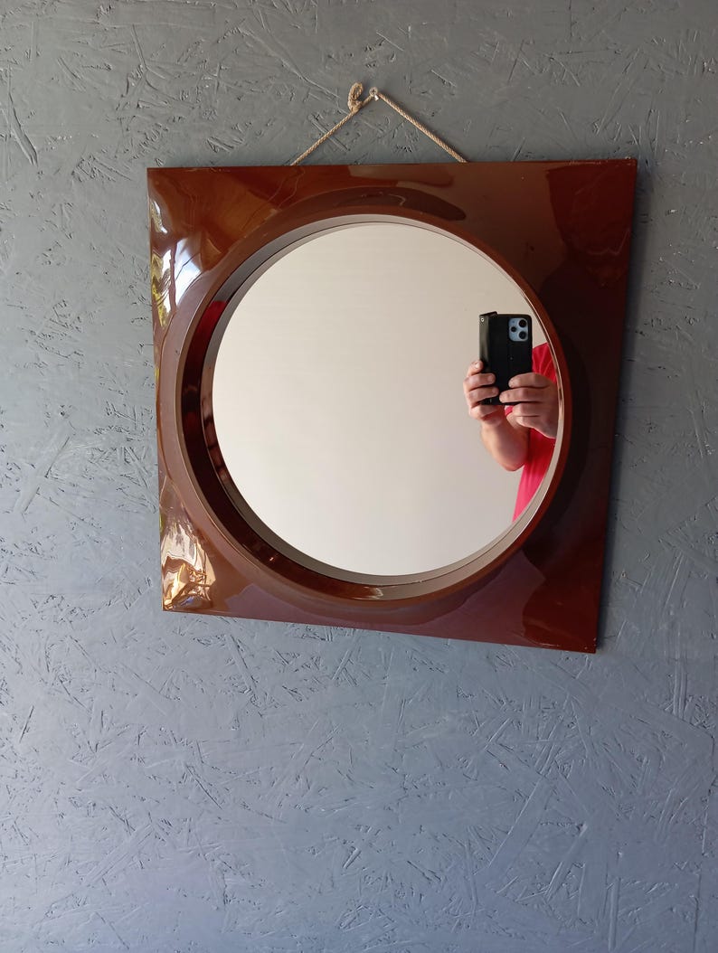 May include: A square wall mirror with a glossy dark brown frame and a circular center glass. The mirror features a simple rope hanger attached to the top edge, allowing it to be mounted securely on a wall surface for display and reflection.