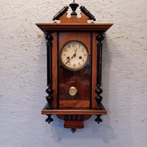 Op de afbeelding: Een vintage houten wandklok met een ronde wijzerplaat en sierlijke details. De klok heeft een donkerhouten frame met zwarte accenten, een glazen paneel en een slinger. De wijzerplaat van de klok heeft zwarte Romeinse cijfers en wijzers.