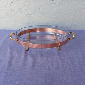 Könnte beinhalten: Eine ovale Glasbackform mit einem kupferfarbenen Metallrahmen und Griffen. Die Form ist leer und steht auf einem kupferfarbenen Metallständer. Die Griffe sind goldfarben. Die Form steht auf einer violetten Oberfläche.