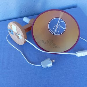Op de afbeelding: Een houten wandlamp met een wit snoer en een beige lampenkap met een bloemenpatroon. De lampenkap heeft een bordeauxrode rand en een wit snoer met een schakelaar.