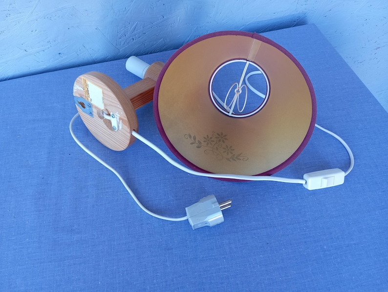 Op de afbeelding: Een houten wandlamp met een wit snoer en een beige lampenkap met een bloemenpatroon. De lampenkap heeft een bordeauxrode rand en een wit snoer met een schakelaar.
