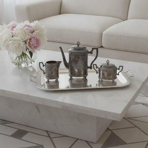 May include: A silver tea set comprising a teapot, a creamer, and a lidded sugar bowl, all arranged on a rectangular silver tray with decorative edges. A clear glass vase filled with white and light pink peony flowers sits nearby.