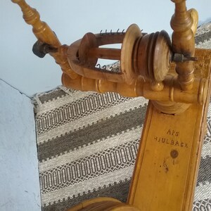 May include: A light brown wooden spinning wheel featuring a flyer assembly with metal hooks and a bobbin. The wooden frame is engraved with the text APS HJULBACK. The structure includes turned wooden uprights and a large circular wheel component.
