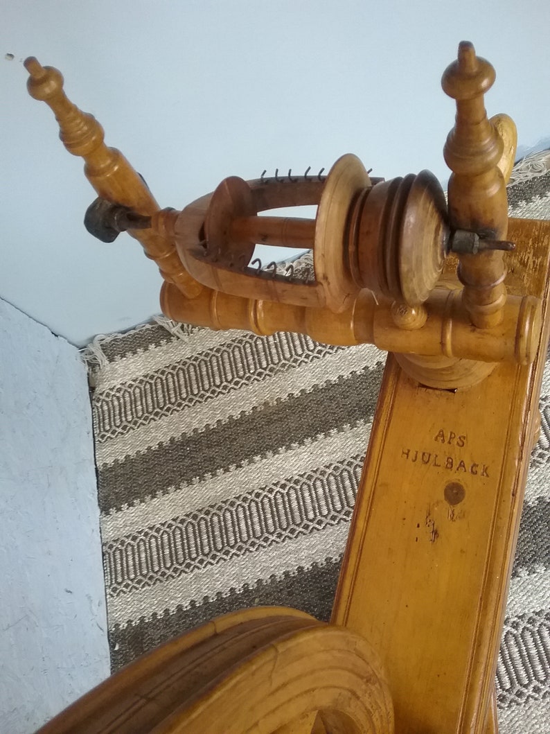 May include: A light brown wooden spinning wheel featuring a flyer assembly with metal hooks and a bobbin. The wooden frame is engraved with the text APS HJULBACK. The structure includes turned wooden uprights and a large circular wheel component.
