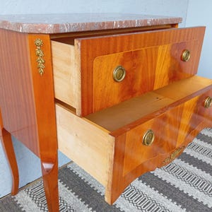 May include: A two-drawer wooden chest with a marble top. The drawers are open, revealing the interior. The chest has brass hardware and decorative accents. The wood has a warm, reddish-brown finish. The chest is on a patterned rug.
