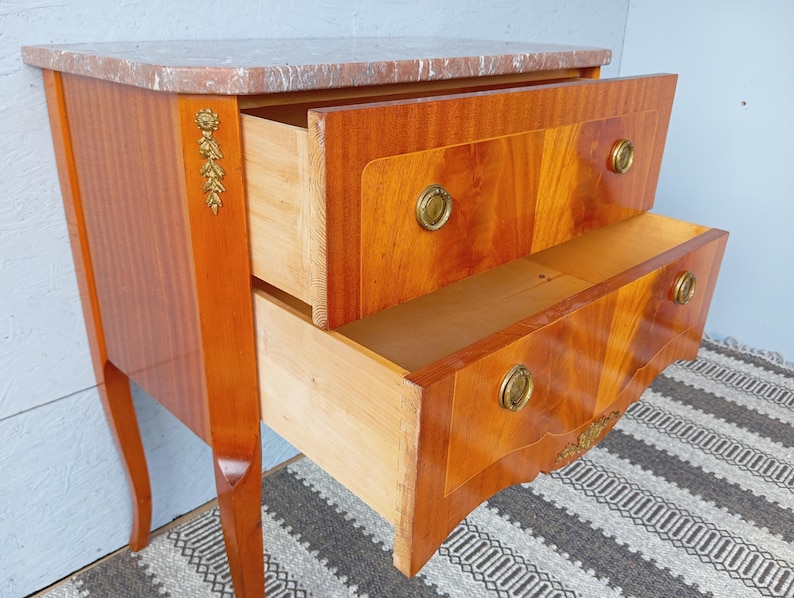 May include: A two-drawer wooden chest with a marble top. The drawers are open, revealing the interior. The chest has brass hardware and decorative accents. The wood has a warm, reddish-brown finish. The chest is on a patterned rug.