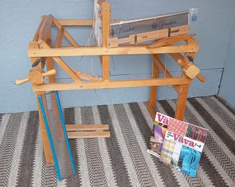 May include: A wooden weaving loom with a light brown finish. The loom has a rectangular frame with various wooden components, including a beater, shuttle, and heddle. A blue and silver tool and magazines are nearby. The loom is set on a patterned rug.