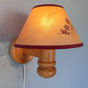 Op de afbeelding: Een houten wandlamp met een cr&egrave;mekleurige lampenkap met een bloemenmotief. De lampenkap heeft een bordeauxrode rand en de wandlamp heeft een natuurlijke houten afwerking.