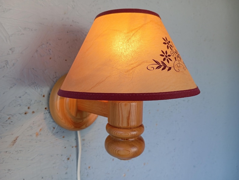 Op de afbeelding: Een houten wandlamp met een cr&egrave;mekleurige lampenkap met een bloemenmotief. De lampenkap heeft een bordeauxrode rand en de wandlamp heeft een natuurlijke houten afwerking.