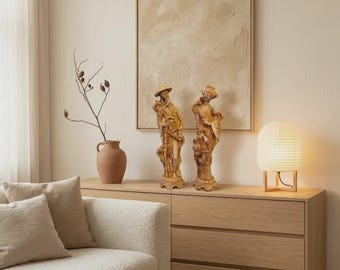 May include: Two beige carved figurines depicting figures in traditional attire, positioned on a light wood dresser alongside a terracotta vase with a dried branch and a round paper lamp with a wooden base, set against a textured beige wall art.