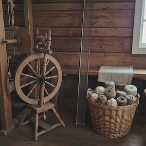 Puede incluir: Una rueca de madera con una rueda circular grande y una base de pedal, situada junto a un marco rectangular alto y estrecho y una gran cesta de mimbre tejida llena de varias bolas de hilo de colores naturales en tonos beige y marrón.