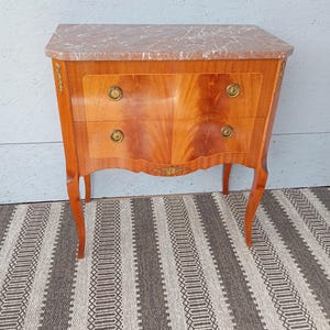 May include: A two-drawer wooden chest with a marble top. The chest has a light brown finish with a curved front and brass hardware. The legs are curved and the chest is on a striped rug.