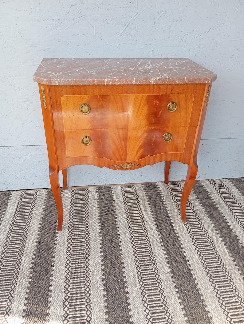 May include: A two-drawer wooden chest with a marble top. The chest has a light brown finish with a curved front and brass hardware. The legs are curved and the chest is on a striped rug.
