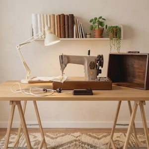 May include: A vintage Singer sewing machine in a beige finish sits on a wooden base with a matching dark wood cover. A white desk lamp illuminates the machine, which is connected to a black foot pedal by a white cord, all placed on a light wood table.