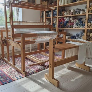 Puede incluir: Un gran telar de madera con una tela blanca natural en proceso, que cuenta con un marco resistente, un banco de madera y una rueda lateral. El telar está colocado sobre una alfombra estampada, con estantes de ovillos de lana de colores al fondo.