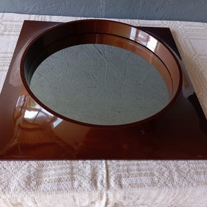 May include: A square wall mirror featuring a glossy, dark brown frame with a circular cutout in the center. The reflective glass surface is set within the deep, polished brown border, creating a distinct contrast between the square outer edge and the round inner mirror section.