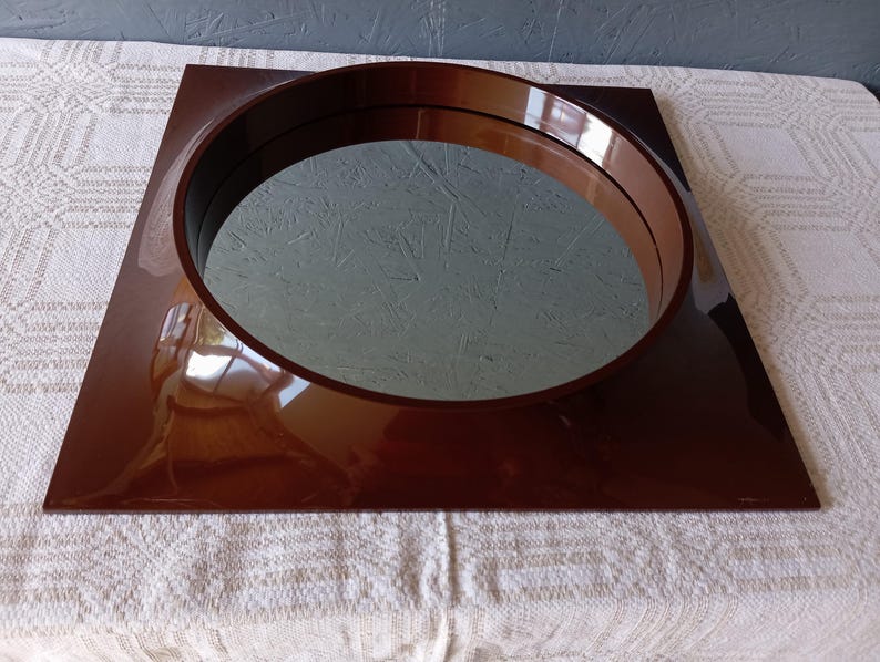 May include: A square wall mirror featuring a glossy, dark brown frame with a circular cutout in the center. The reflective glass surface is set within the deep, polished brown border, creating a distinct contrast between the square outer edge and the round inner mirror section.