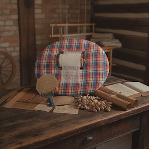 Può includere: Un cuscino rotondo per merletti con motivo a quadri rosso, blu e bianco. Il cuscino sostiene un telaio centrale in legno con una striscia di pizzo bianco. Vicino ci sono fuselli in legno, cartamodelli, un piccolo attrezzo e un libro.
