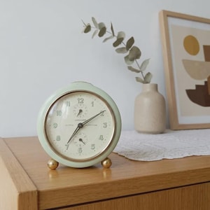 May include: A round, pale mint green analog alarm clock with a cream-colored face, gold-toned hands, and gold-toned spherical feet. The clock face features black numerals and the text Junghans and Bivox Repetition printed in the center.