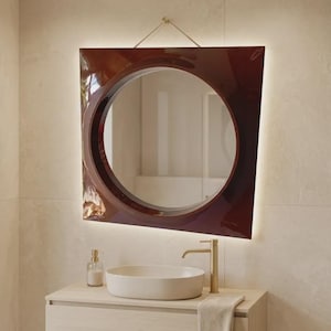 May include: A wall-mounted mirror with a glossy dark red, trapezoidal frame and a circular center. The mirror features integrated backlighting, a gold-toned faucet, a white oval vessel sink, and a light wood vanity cabinet with a hanging towel.