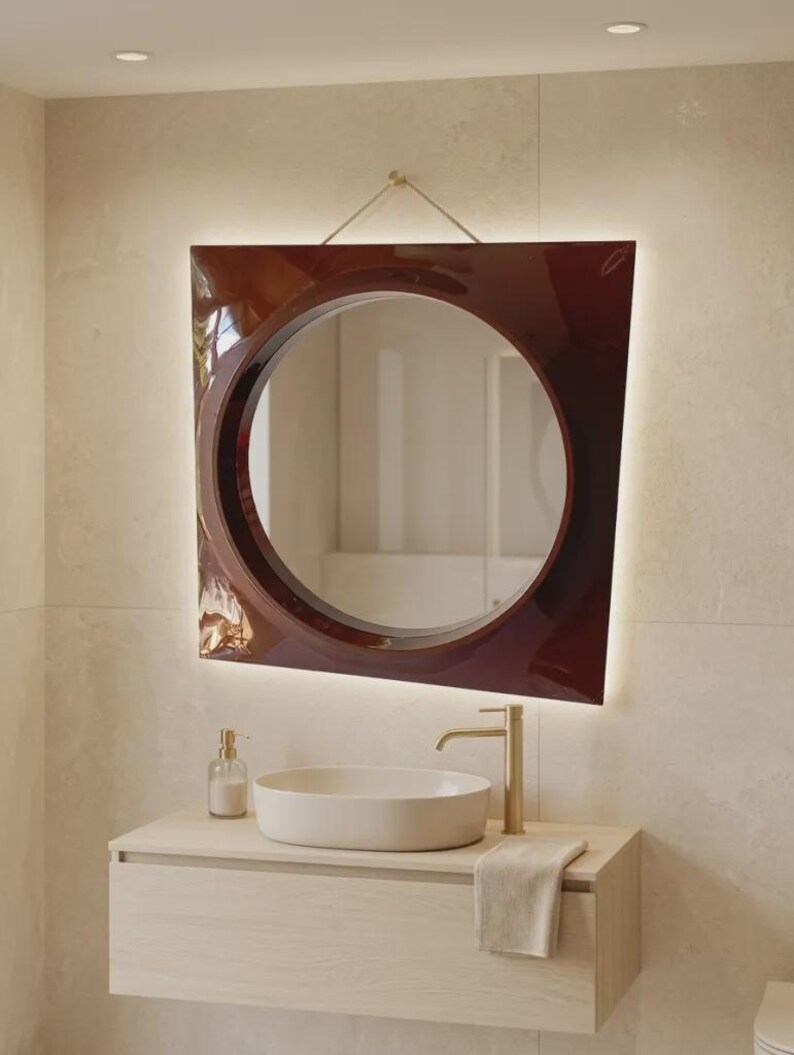 May include: A wall-mounted mirror with a glossy dark red, trapezoidal frame and a circular center. The mirror features integrated backlighting, a gold-toned faucet, a white oval vessel sink, and a light wood vanity cabinet with a hanging towel.