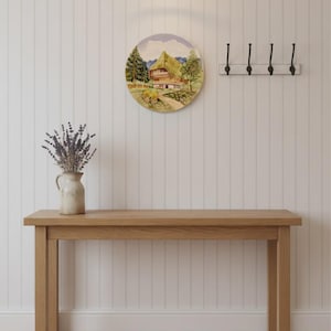 Puede incluir: Un plato decorativo redondo de cerámica con un paisaje pintado de una casa de madera tradicional entre árboles verdes y montañas. El plato se exhibe en una pared con paneles blancos sobre una mesa consola de madera con un jarrón de lavanda.