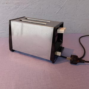 May include: A vintage two-slice toaster with a silver ribbed front and black accents. The toaster has a white lever and a black power cord with a plug. The appliance is on a pink surface.