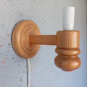 Op de afbeelding: Een houten wandlamp met een witte lampfitting. De lamp is gemaakt van natuurlijk hout met een lichtbruine afwerking. De lamp heeft een ronde basis en een gebogen arm die naar de zijkant uitsteekt. De lampfitting is bevestigd aan het uiteinde van de arm.