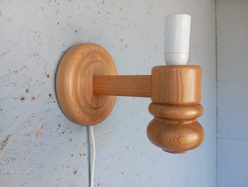 Op de afbeelding: Een houten wandlamp met een witte lampfitting. De lamp is gemaakt van natuurlijk hout met een lichtbruine afwerking. De lamp heeft een ronde basis en een gebogen arm die naar de zijkant uitsteekt. De lampfitting is bevestigd aan het uiteinde van de arm.