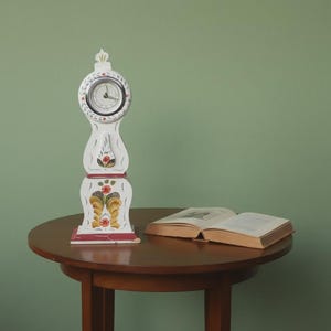May include: A white decorative clock with a round face and floral patterns. It stands on a small, round, brown table beside an open book. The clock's base features red accents. The clock is a decorative item.