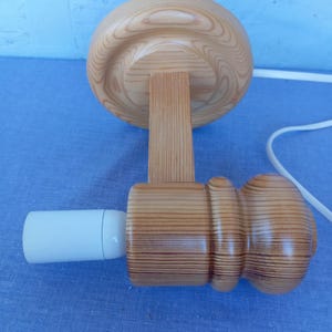 Op de afbeelding: Een houten wandlamp met een ronde, lichtbruine basis en een cilindrische, lichtbruine kap. De lamp heeft een witte fitting en een wit snoer.