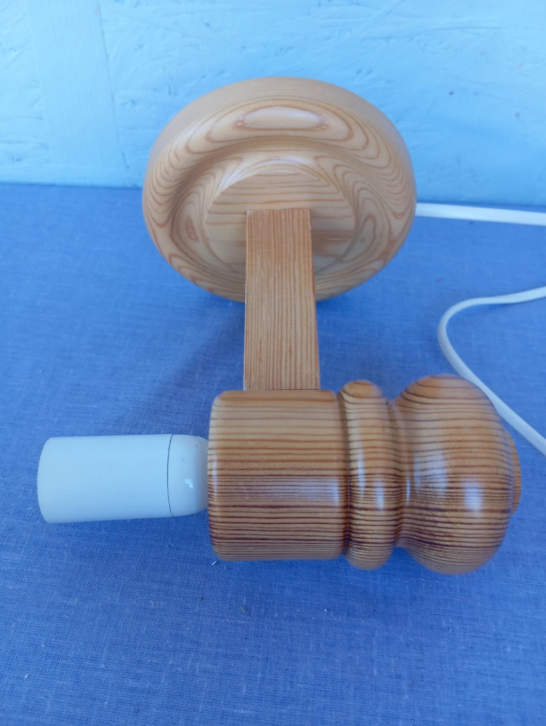 Op de afbeelding: Een houten wandlamp met een ronde, lichtbruine basis en een cilindrische, lichtbruine kap. De lamp heeft een witte fitting en een wit snoer.