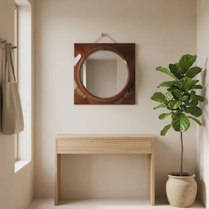 May include: A square wall mirror with a glossy dark brown frame and a circular center glass, mounted with a silver cord. The mirror is positioned above a light-toned wooden console table with a single drawer and straight legs.