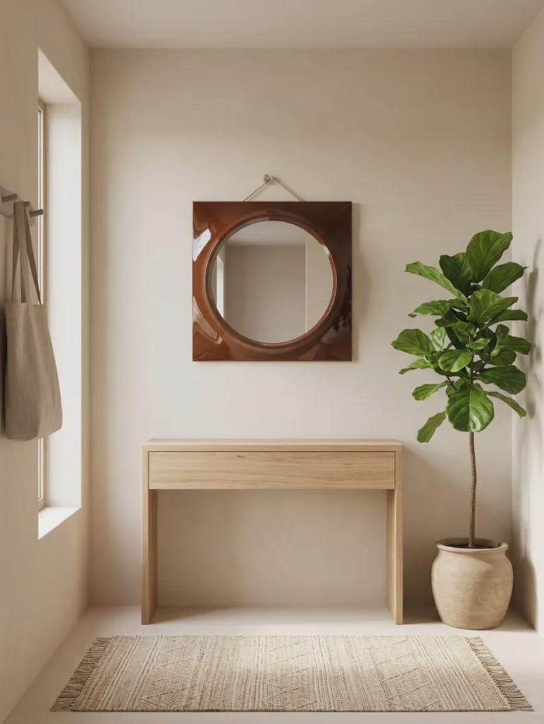 May include: A square wall mirror with a glossy dark brown frame and a circular center glass, mounted with a silver cord. The mirror is positioned above a light-toned wooden console table with a single drawer and straight legs.
